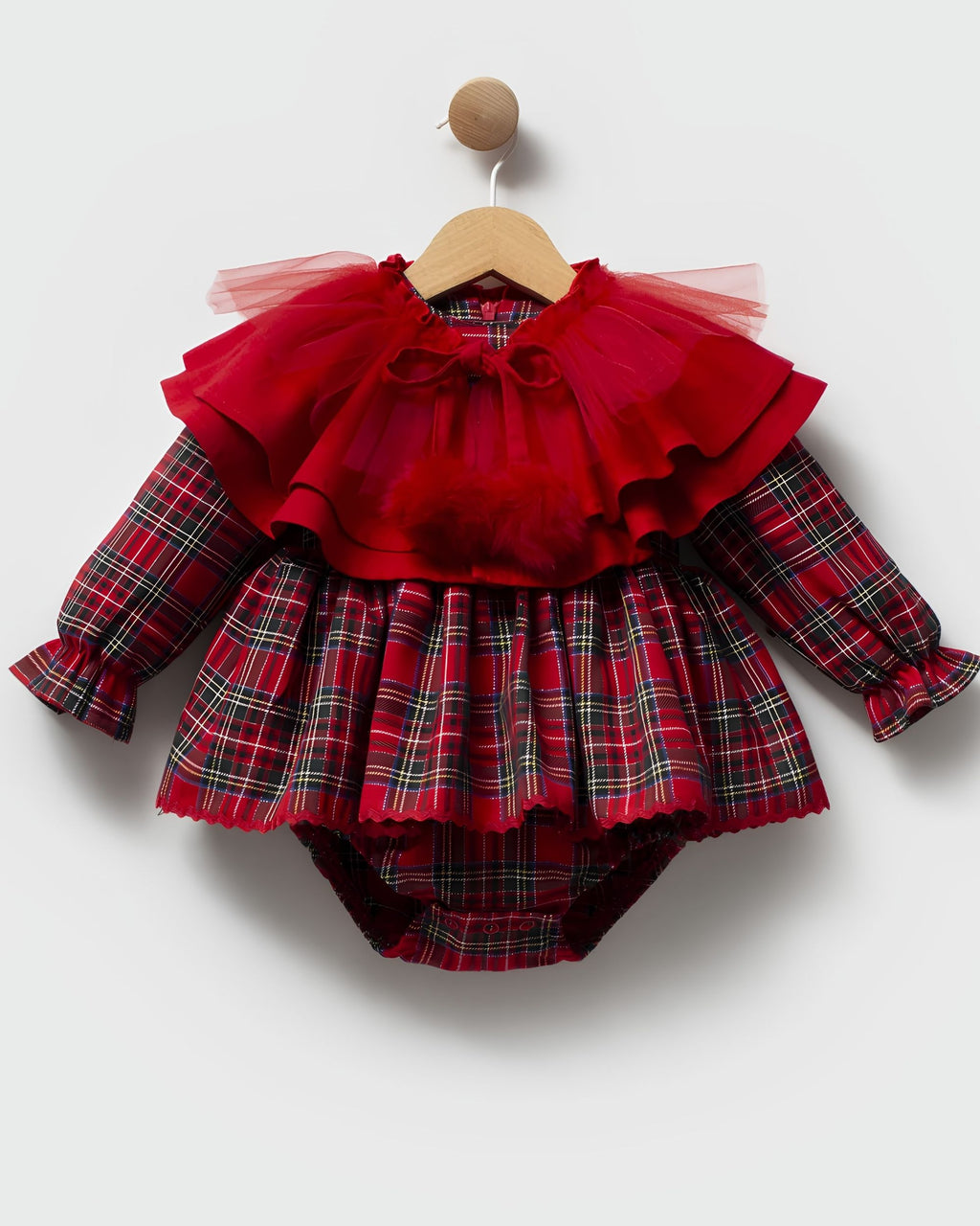 A baby girl's long-sleeve romper in red plaid with a layered red tulle ruffle collar and bow, hanging on a wooden hanger against a light gray background.