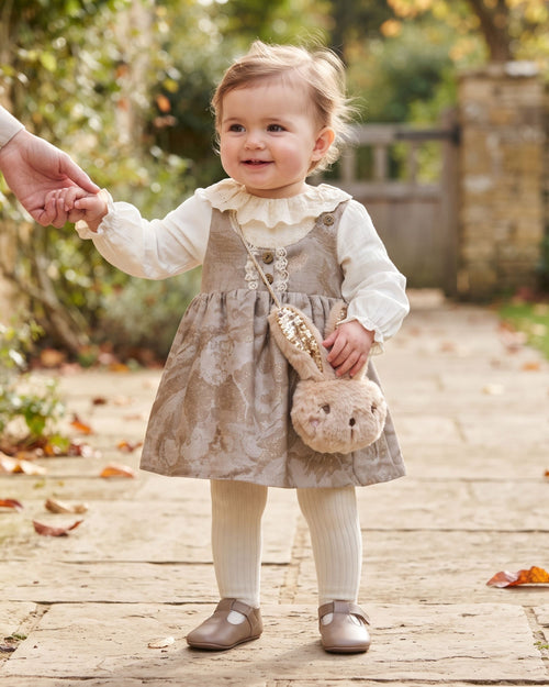 Baby girl wearing a taupe pinafore dress and carrying a plush bunny bag