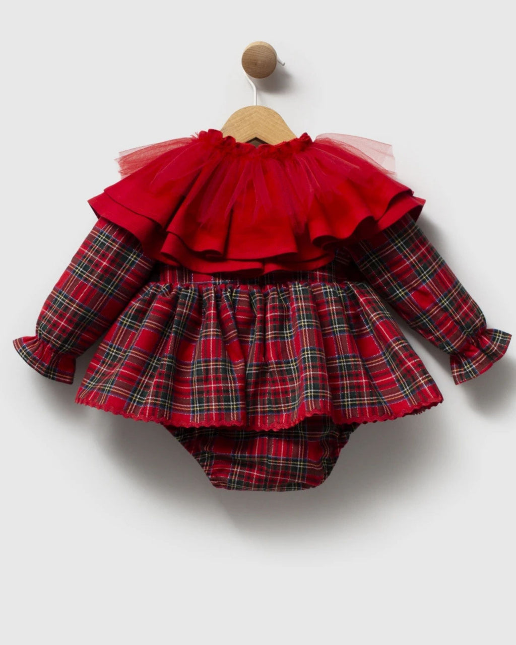 A baby girl's long-sleeve romper in a red and green plaid fabric, featuring a layered red tulle ruffle collar and a matching ruffled hem, hanging on a wooden hanger against a light gray background.